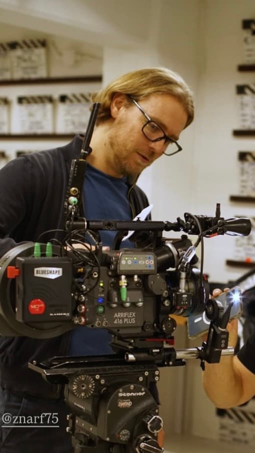 Blonde man with glasses inspecting a professional Arriflex film camera in a studio setting.