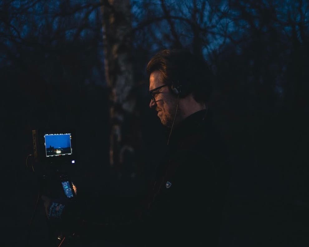 Man with headphones and glasses looks at a glowing camera monitor in a dark forest.