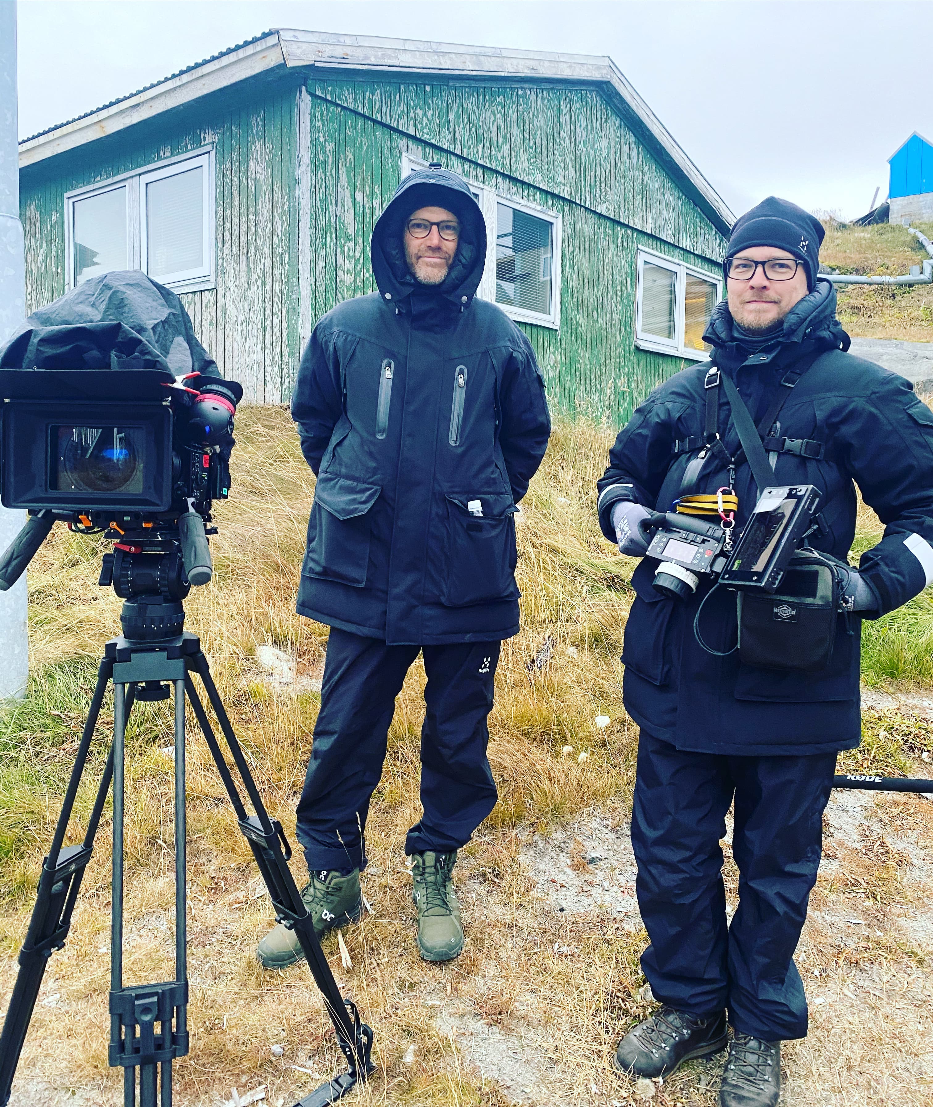 Two men in winter gear stand with professional cameras outside a weathered green wooden building.