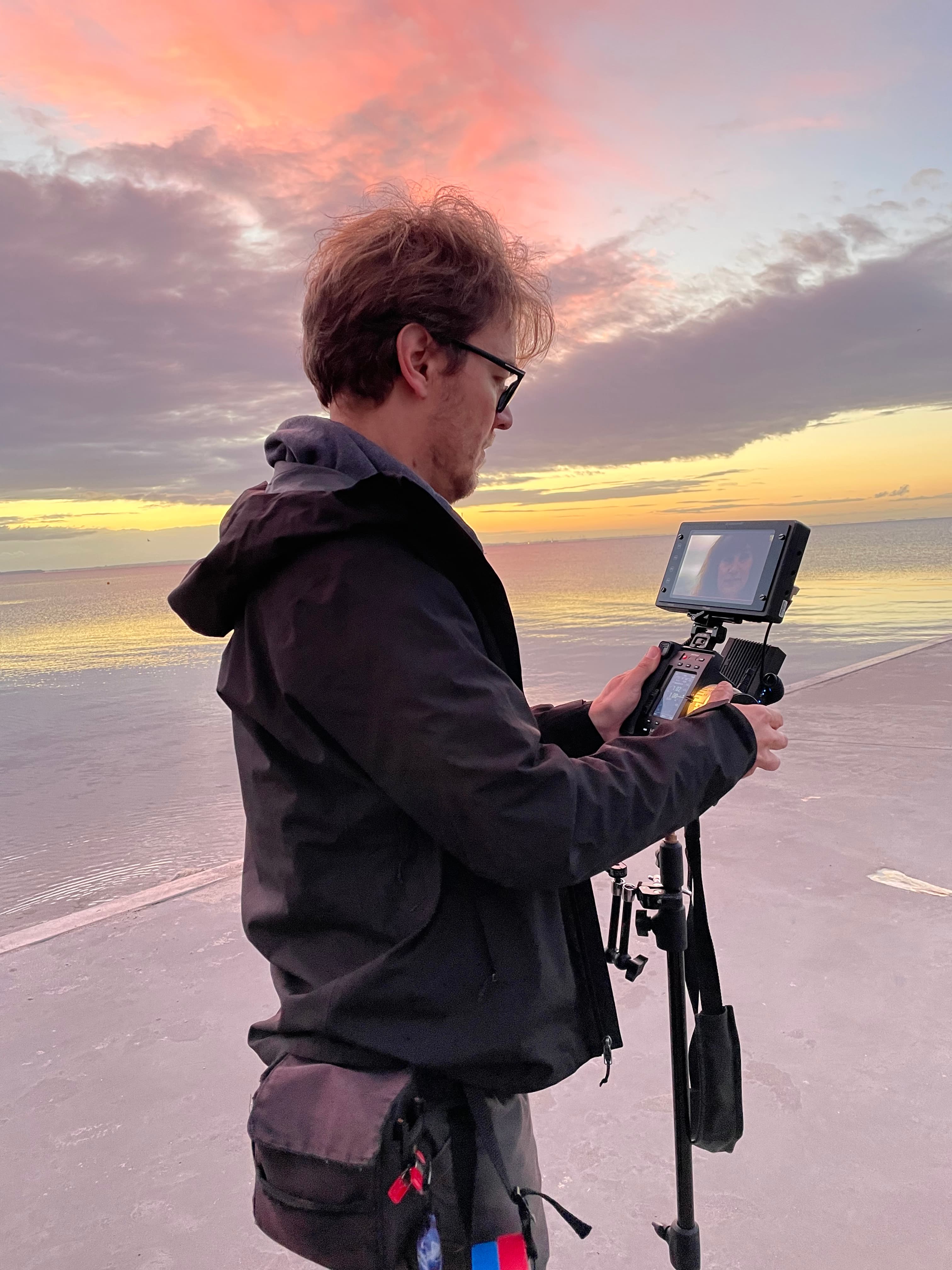 Videographer filming a colorful sunset over the ocean with a professional camera and monitor.